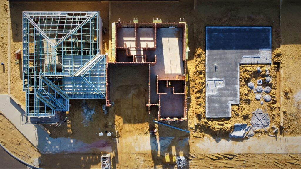 overhead/satellite/drone image of three houses in various stages of construction against a sandy dirt background, the house on the left has its roof struts framed out, the middle one just has some walls, the one on the right is only the concrete slab